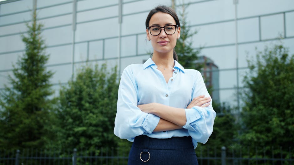 Confident businesswoman standing with arms crossed, wearing glasses, outside office building.